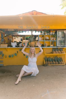 Photo by Nina Hill Happy woman in white dress at tropical fruit stand raising hands with pineapple.