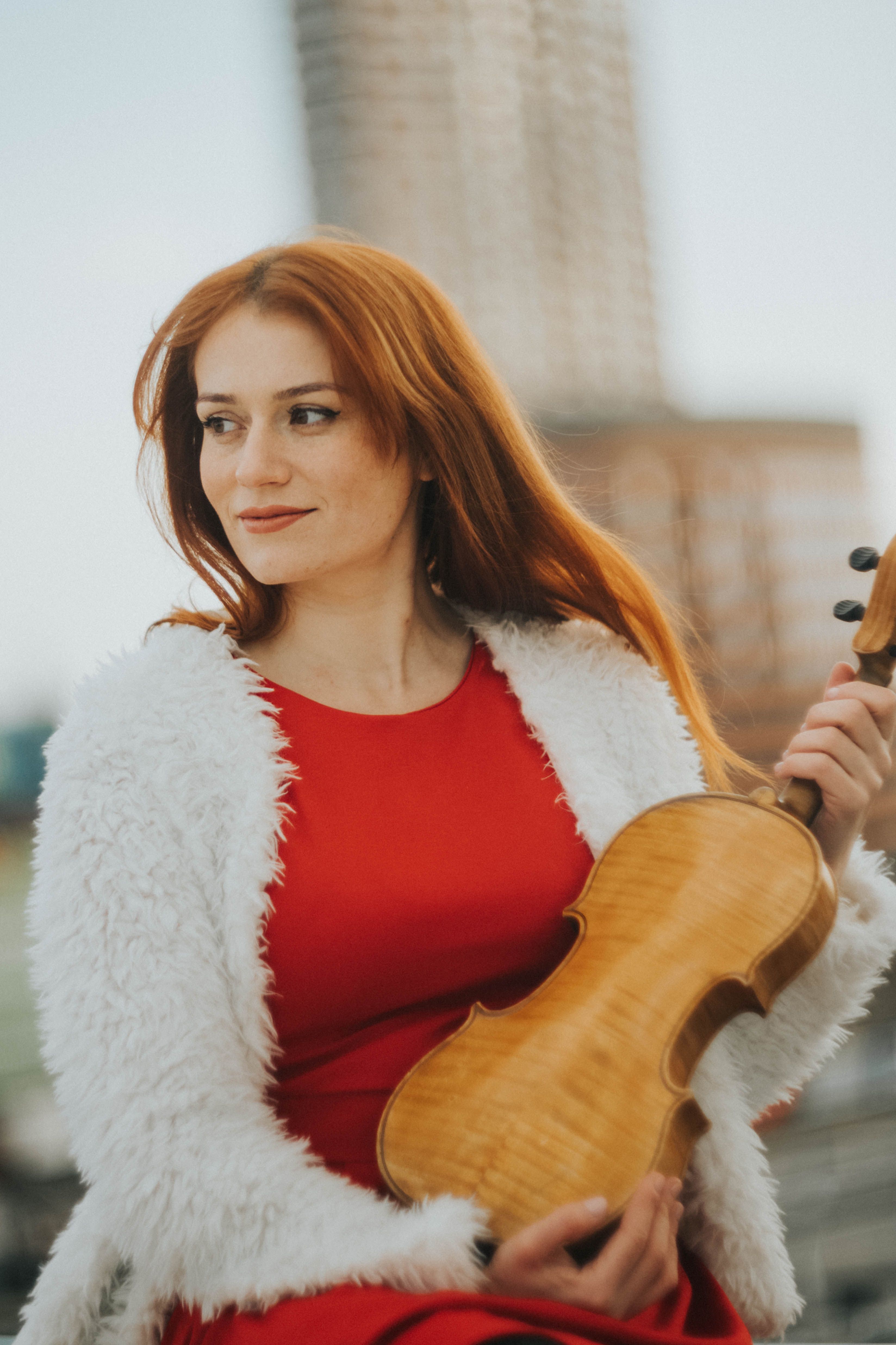 A woman with red hair in a vibrant red dress holding a violin against an urban background.