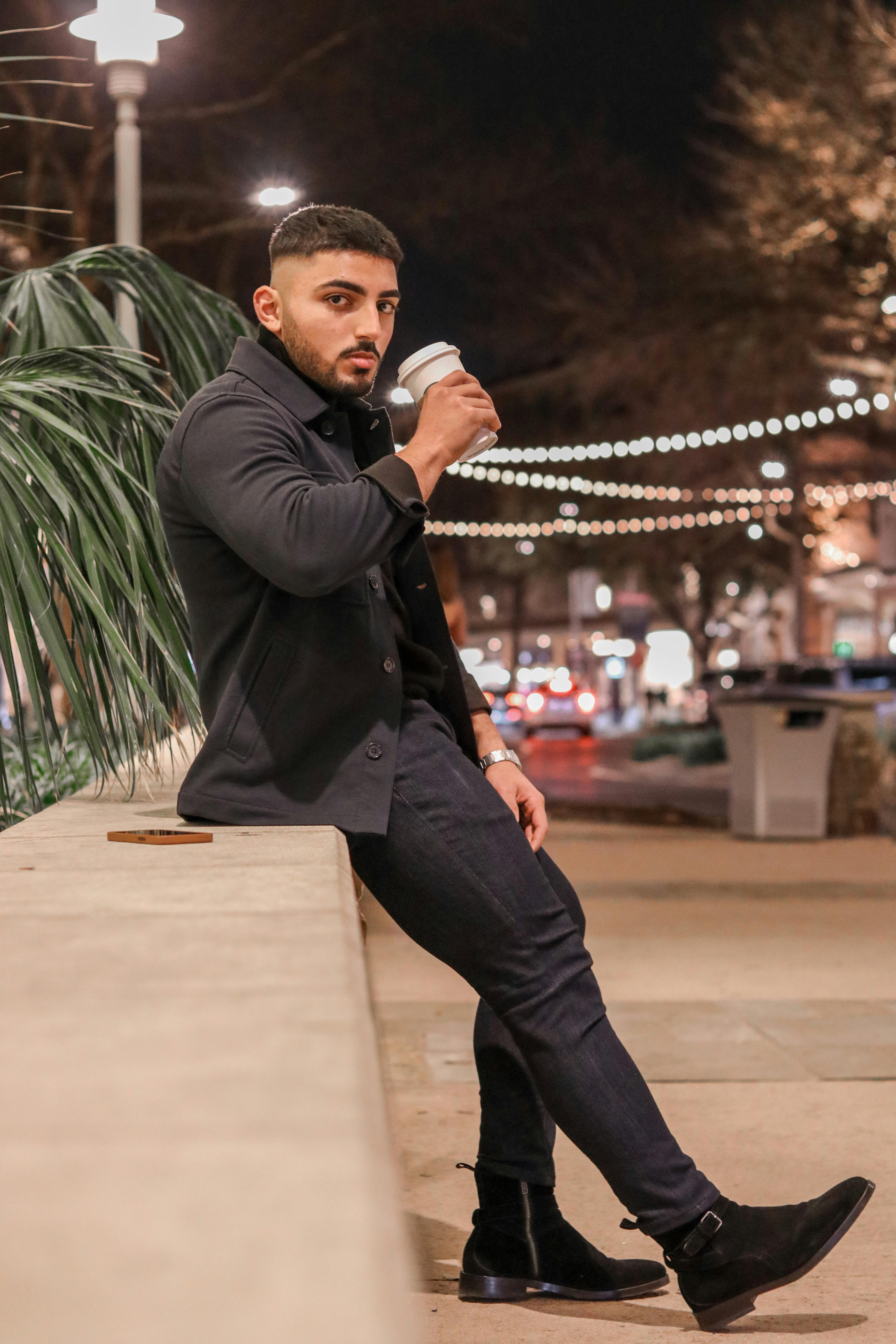 A man sits outdoors at night enjoying a coffee amidst city lights.