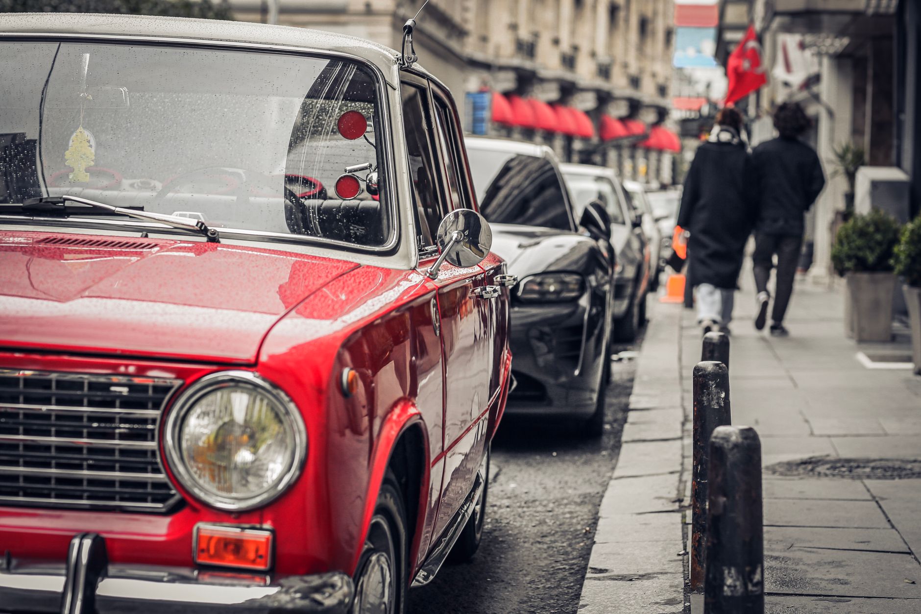 Red vintage car parked on a rainy street in Istanbul, with urban scenery and pedestrians in the background.