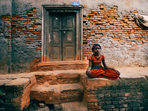 Free A young girl in red attire sits by an old brick facade with a wooden door, creating a rustic outdoor scene. Stock Photo