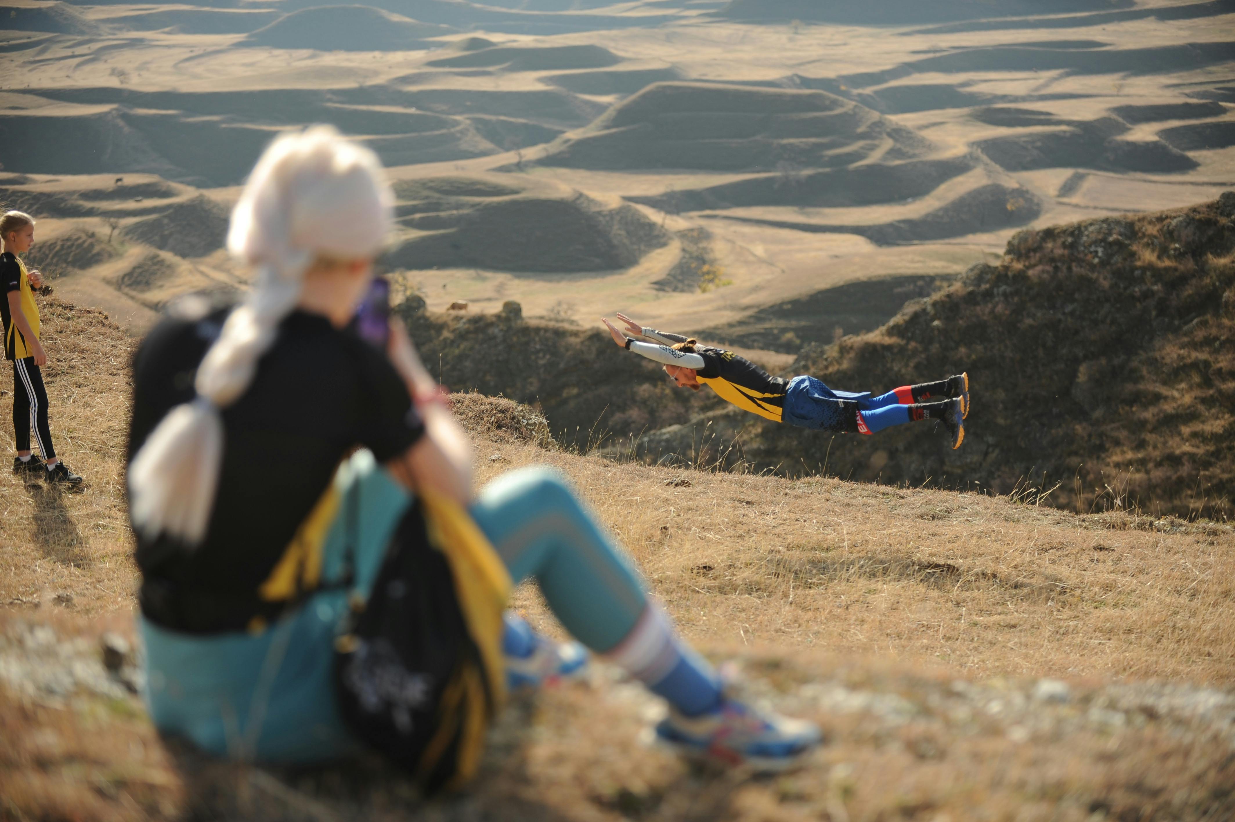 A daring individual performs a leap in a rugged mountain landscape as others observe.