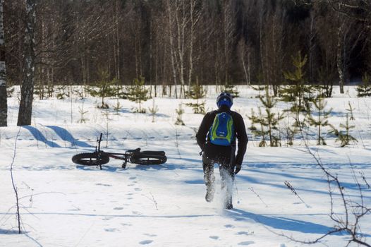 Man with backpack walking in snowy forest near bicycle, embracing winter adventure.