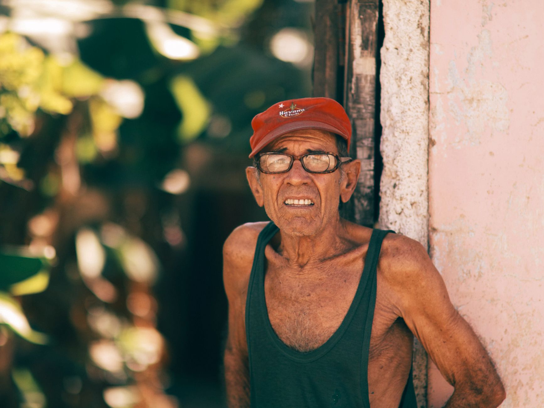 Portrait of a senior man wearing a red cap and eyeglasses, standing outdoors in sunlight.