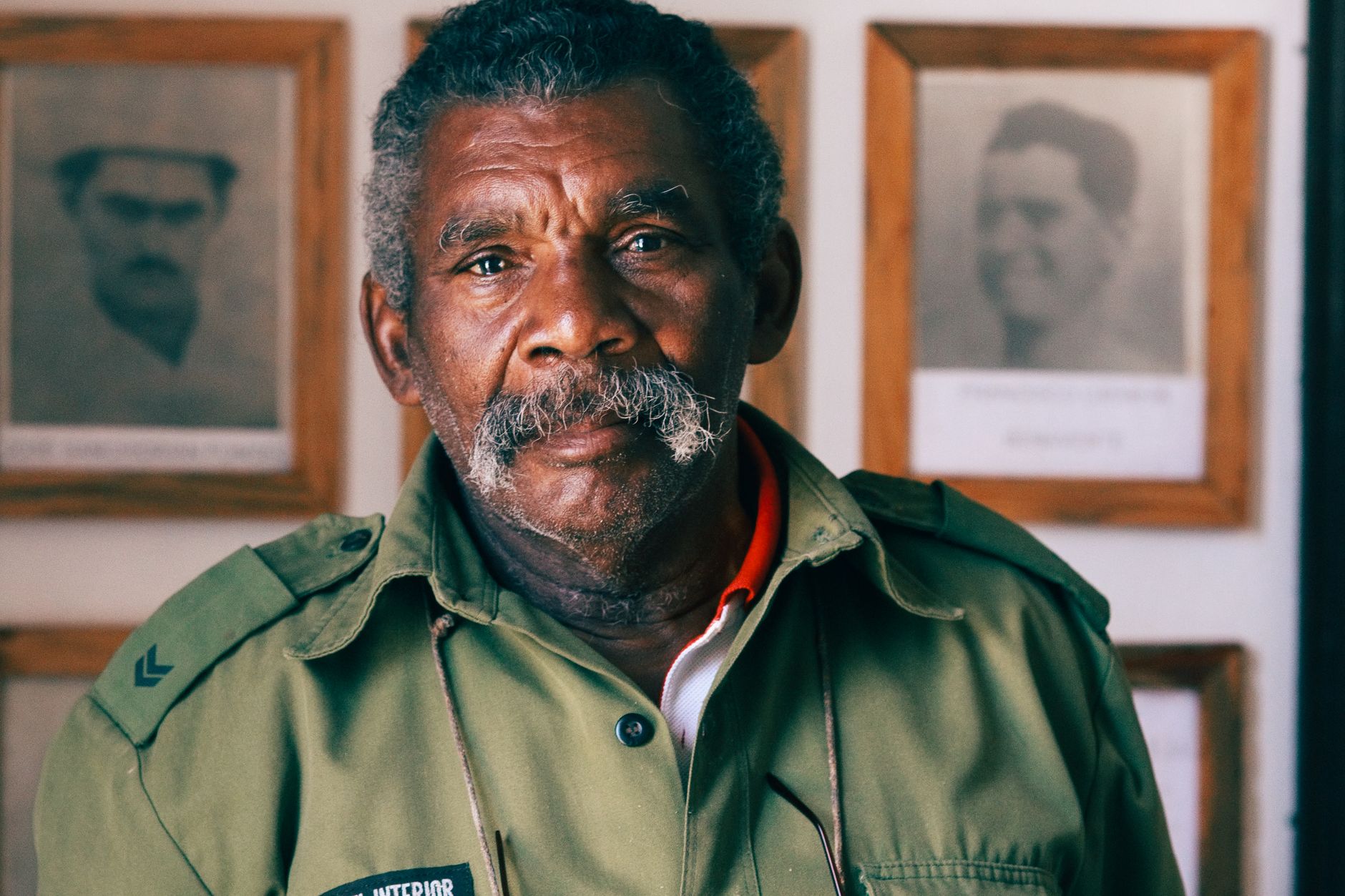 Elderly man with mustache in military uniform, framed photos in background.