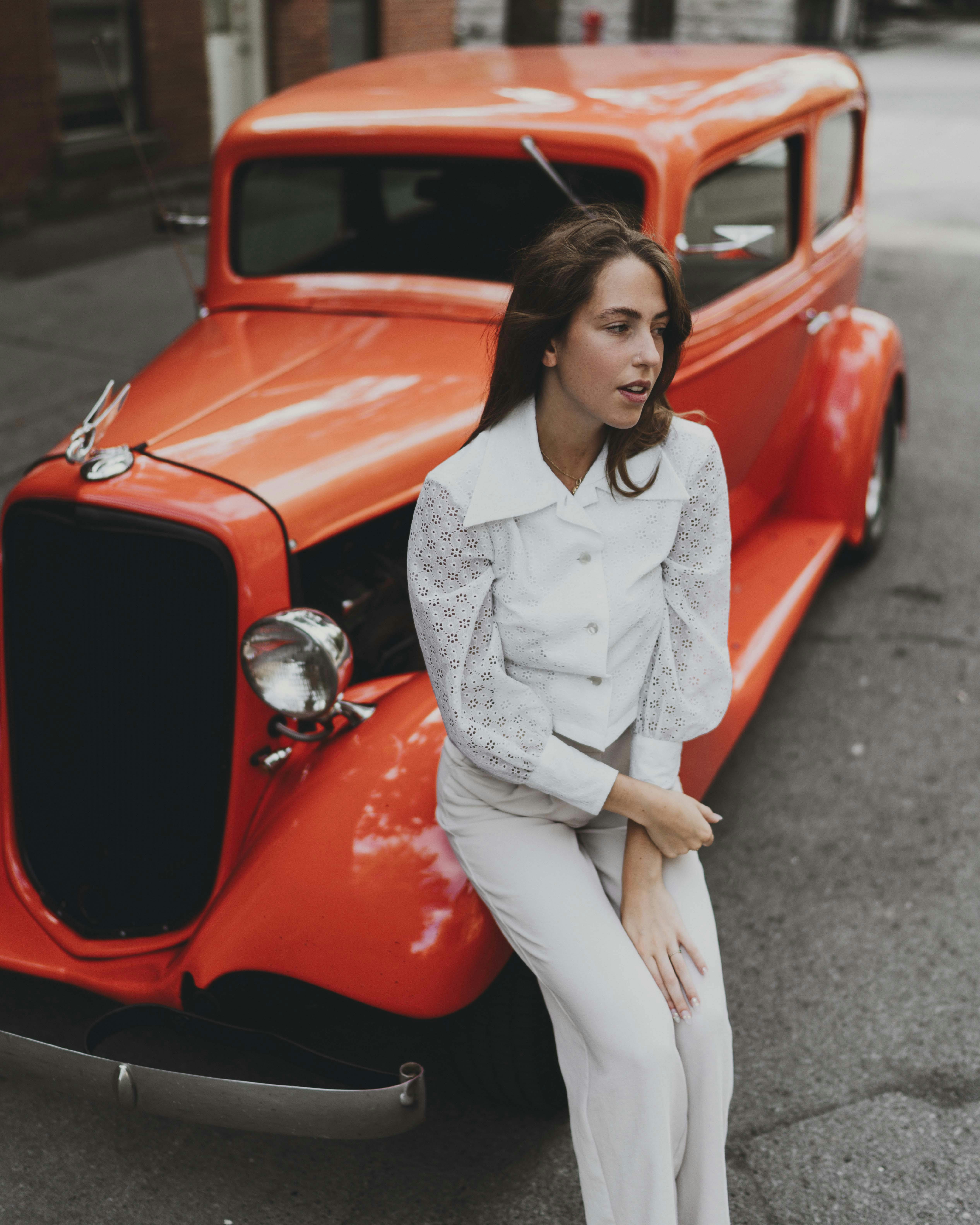 Stylish woman in white, sitting by a vibrant orange vintage car on a city street.
