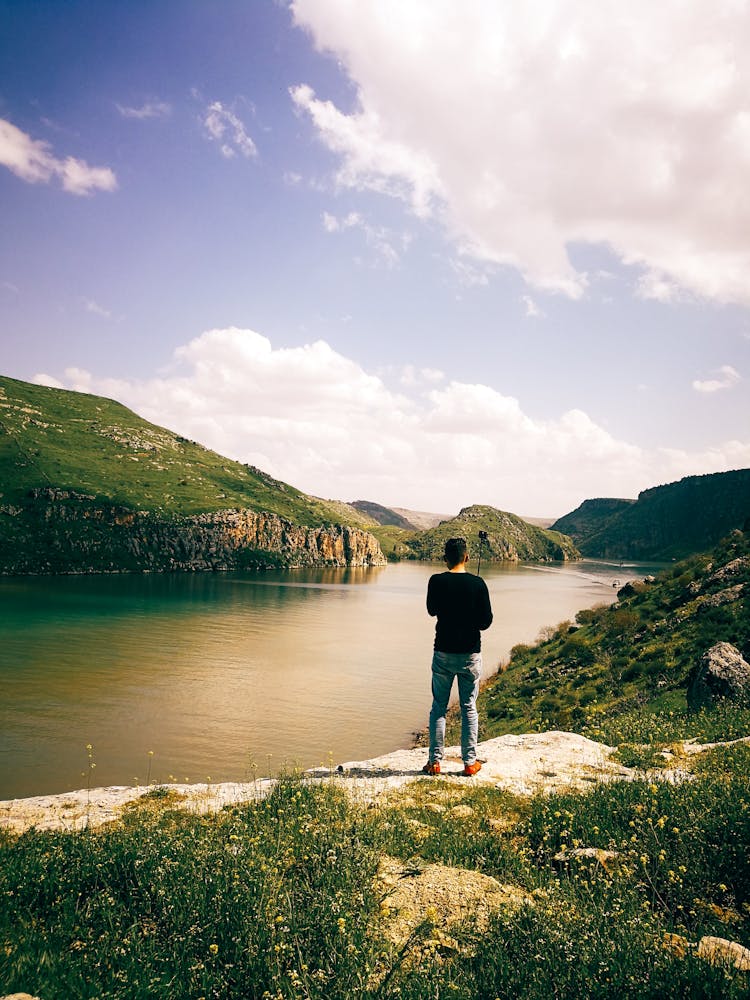 Back View Of A Man Standing On The River Bank In Mountains 