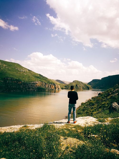 Free A man stands on a riverside cliff, enjoying the stunning valley landscape under clear skies. Stock Photo