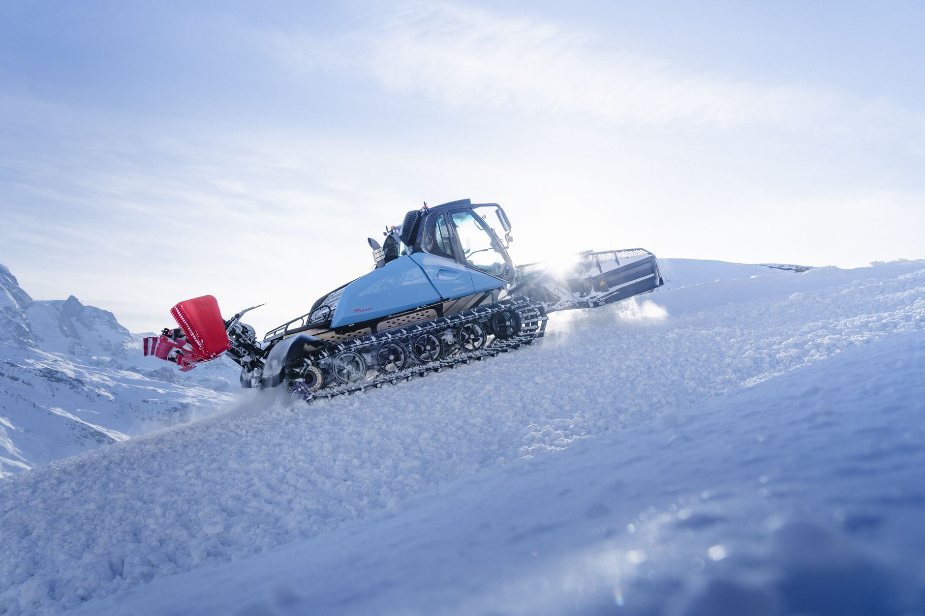 A snow groomer efficiently moves across a snowy landscape in Zermatt, Switzerland under a bright winter sky.