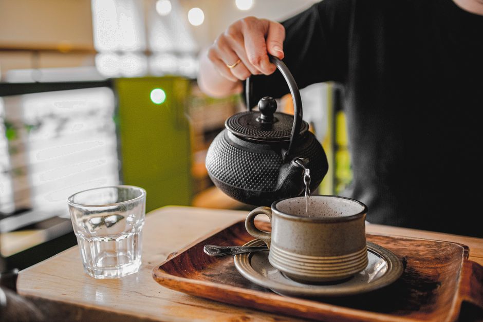 A person pours hot tea from a cast iron teapot into a cup on a wooden tray.