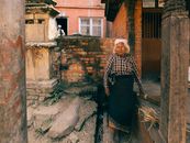 Elderly woman stands by a rustic brick wall holding a broom in a Nepalese street.
