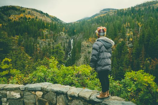 A child in winter clothing admires a waterfall amidst lush, green mountain scenery.