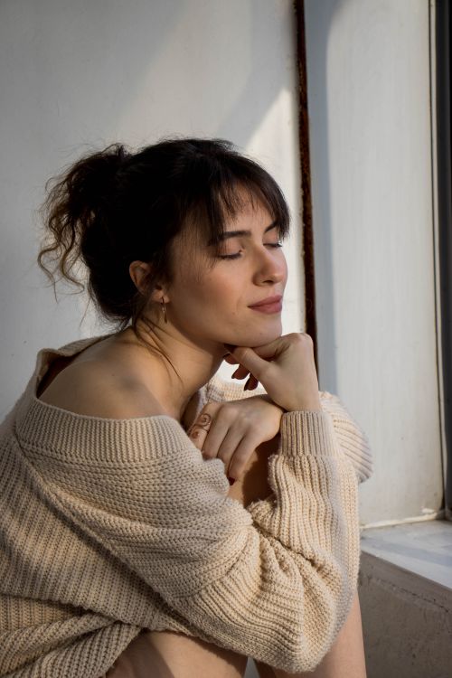 Free A serene woman sits by a window, eyes closed, wearing a knitted sweater in soft light. Stock Photo
