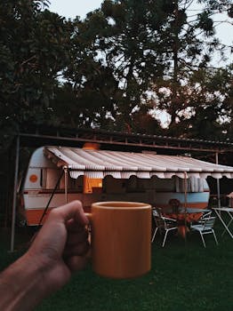 日落时分，一只手拿着咖啡杯，旁边停着一辆拖车，构成了一幅宁静的露营场景