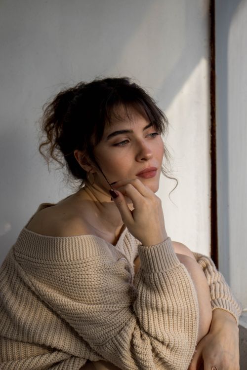 Free A thoughtful woman in a knitted sweater sits by a window, basking in sunlight. Stock Photo