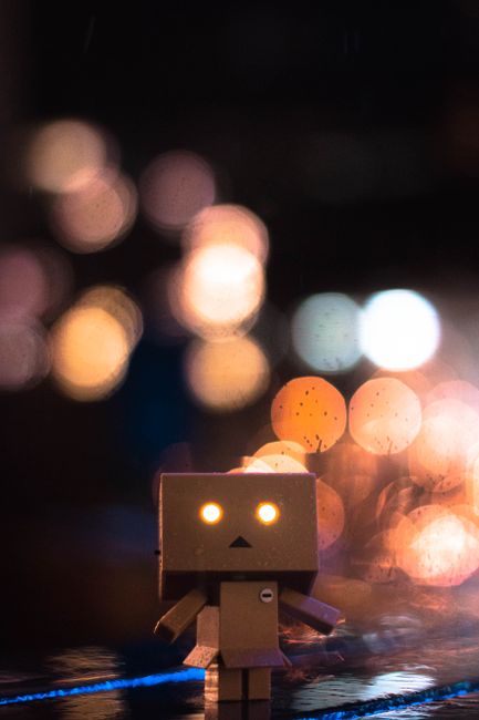 Photo by Aaron Ng A captivating cardboard robot toy set against vibrant bokeh lights, creating a whimsical night scene.