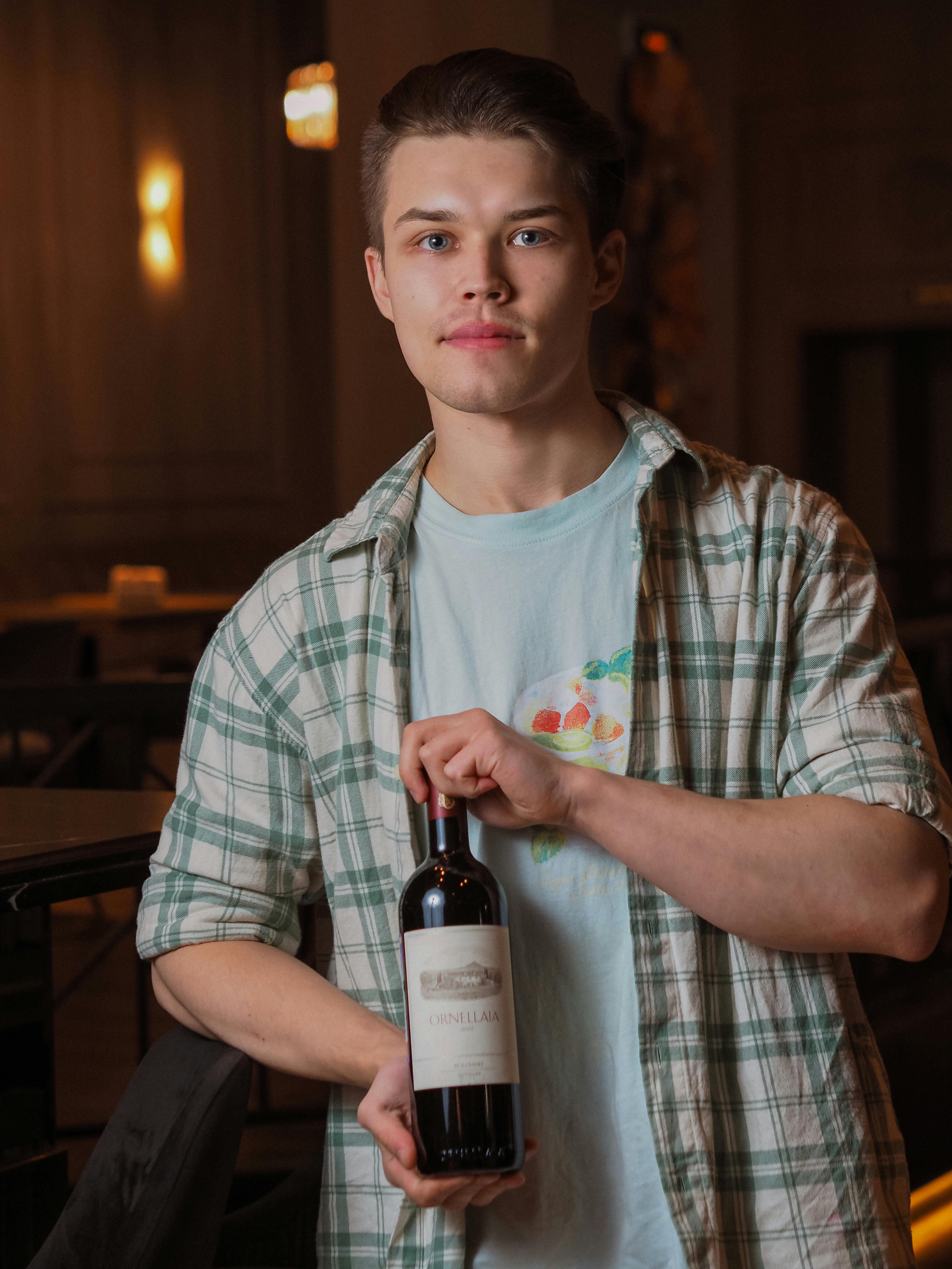 A young man holds a wine bottle indoors, wearing a plaid shirt.