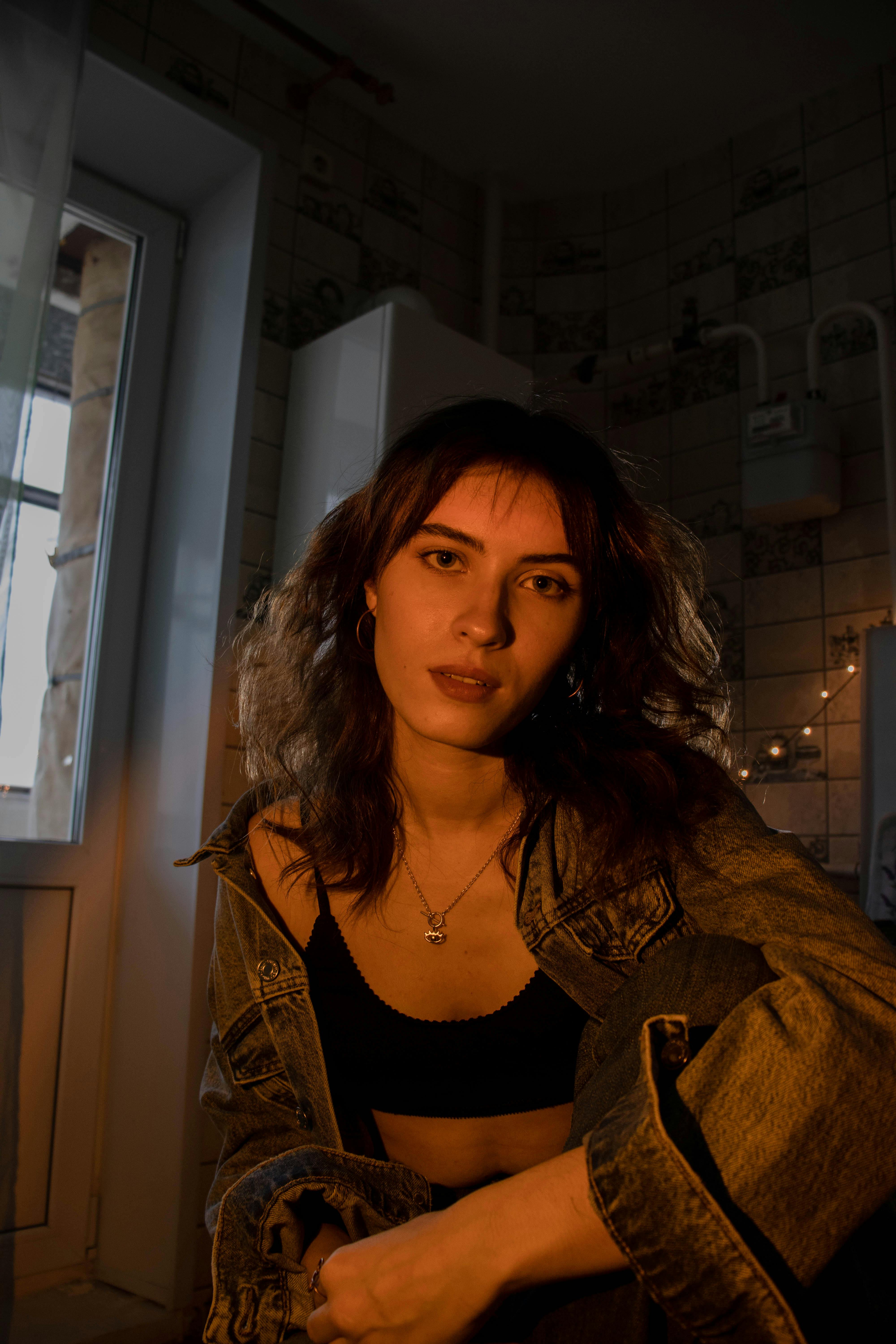 A woman sitting indoors wearing a denim jacket, illuminated with moody lighting, creating a warm and casual atmosphere.