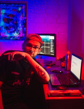 A man sitting at a desk with illuminated screens in a neon-lit room.
