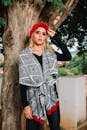 一位戴着红帽子、披着图案披肩的年轻女子在户外的一棵树旁摆姿势
