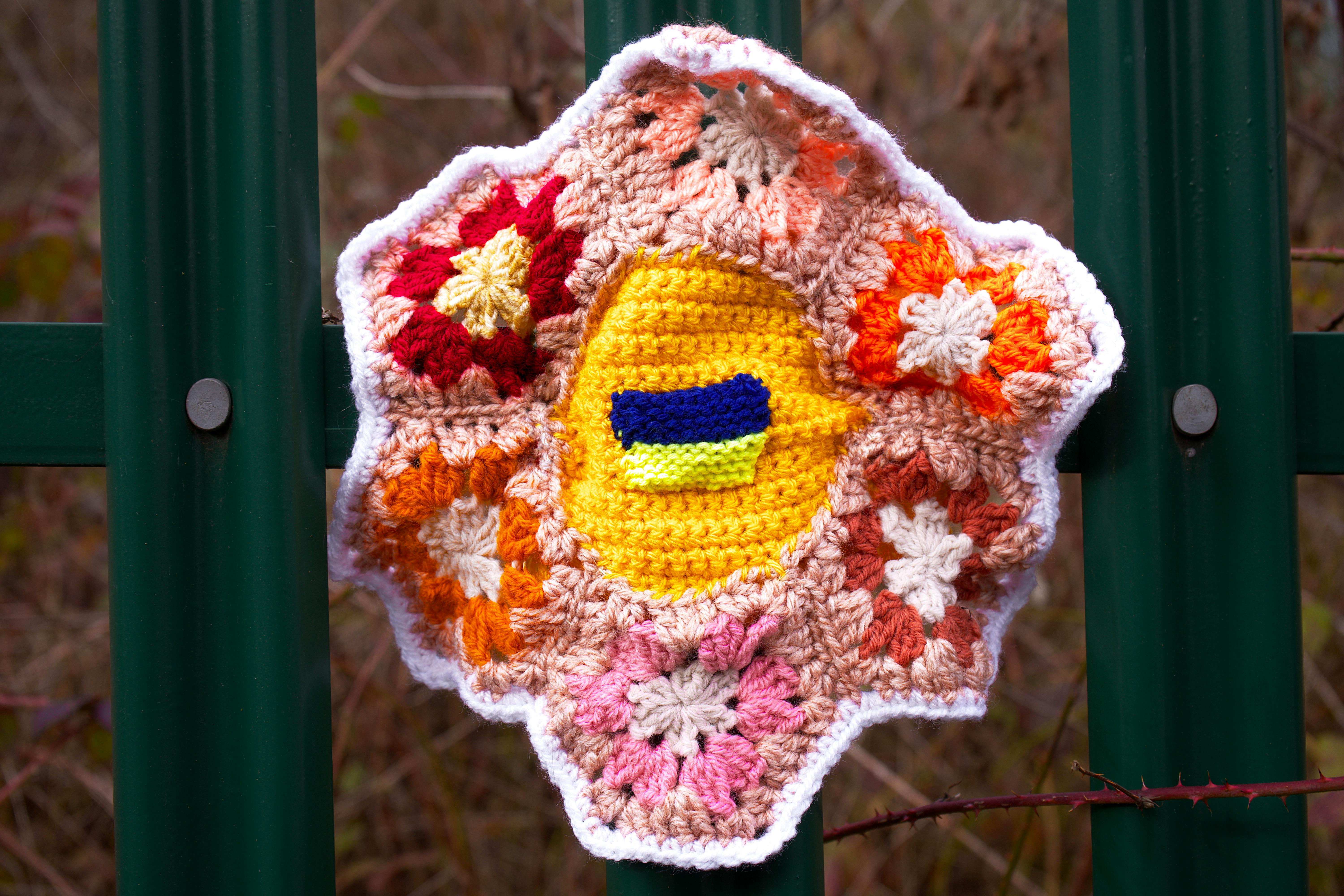 Colorful handmade crochet flower attached to a green fence outdoors in Knebworth, UK related to buy detailed crochet patterns