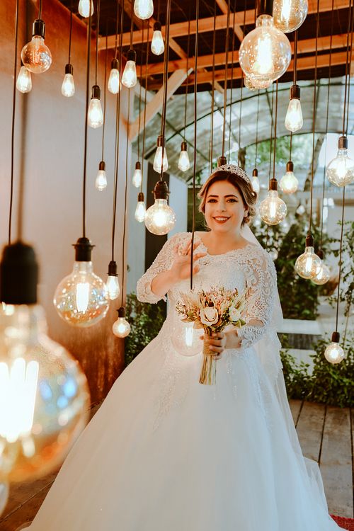 Free Bride in a lace gown holding a bouquet, surrounded by hanging bulbs, creating a magical ambiance. Stock Photo