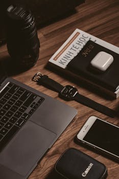 A minimalist desk setup featuring a laptop, smartphone, camera lens, and smart watch.