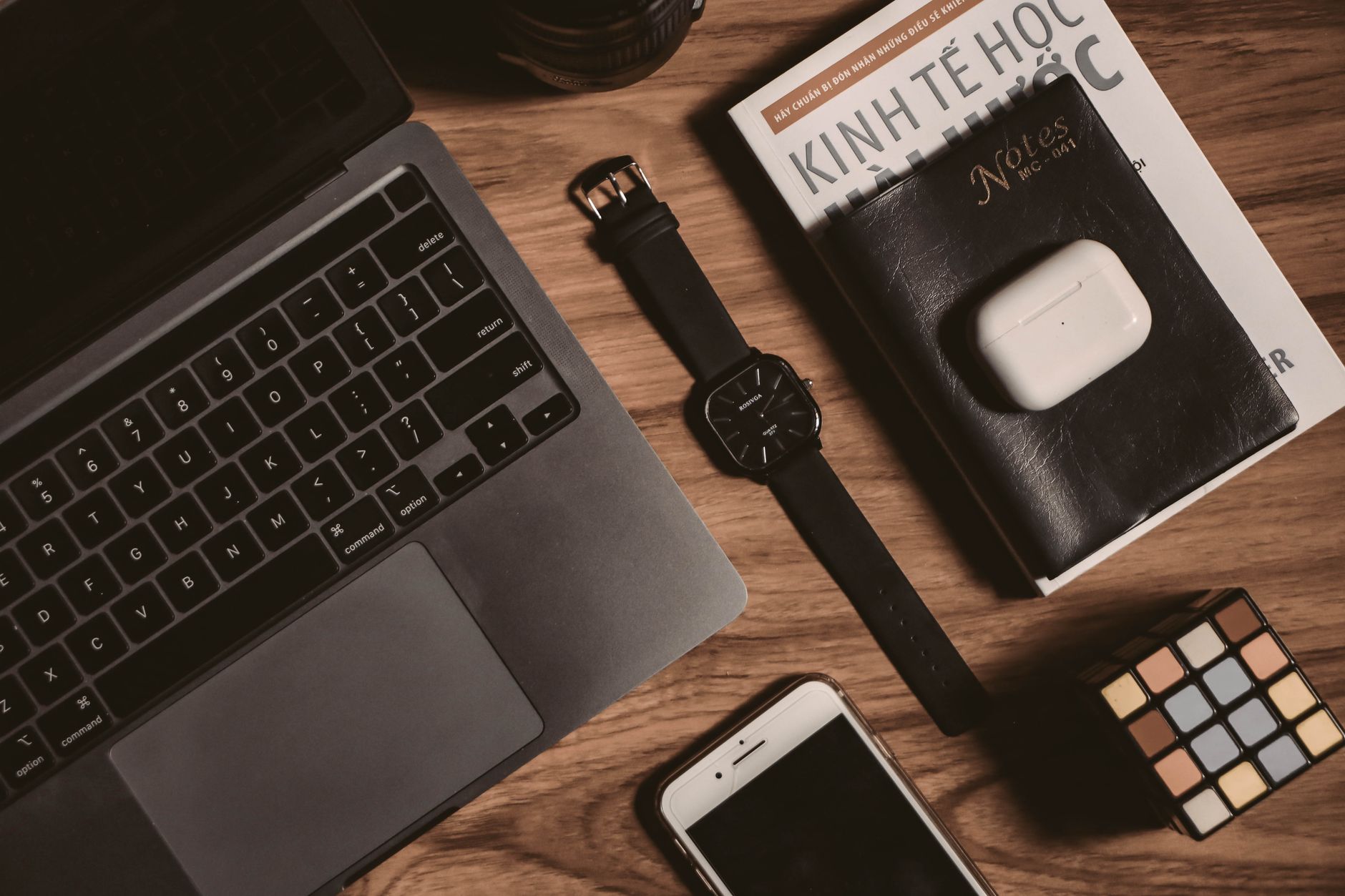 Flatlay of tech gadgets with laptop, smartphone, Rubik's cube, book, watch, and AirPods on a wooden table.
