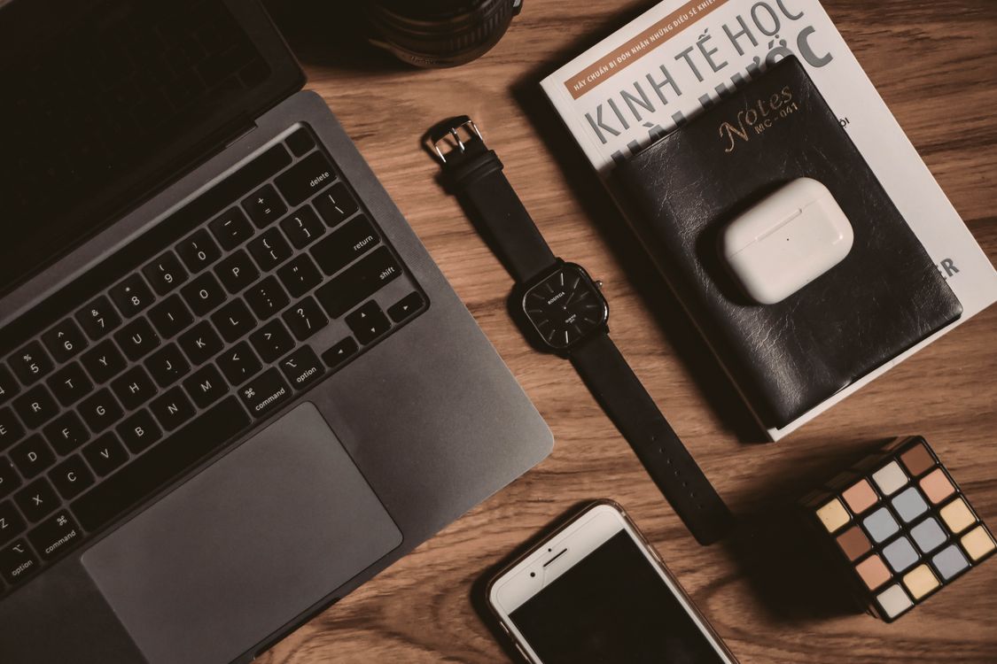Flat lay of tech gadgets including a laptop, smartphone, book, watch, and AirPods on a wooden table