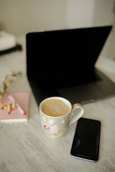 A warm workspace scene featuring a laptop, smartphone, and a floral mug of coffee.