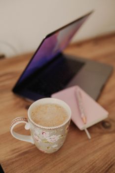 A floral mug of coffee sits beside a laptop and notebook on a wooden desk, creating a cozy workspace.