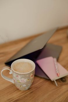 A warm and inviting workspace featuring a floral mug, notebook, and laptop on a wooden surface.