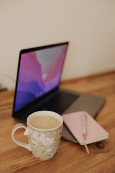 A warm, inviting workspace featuring a laptop, floral mug of coffee, and notebook.