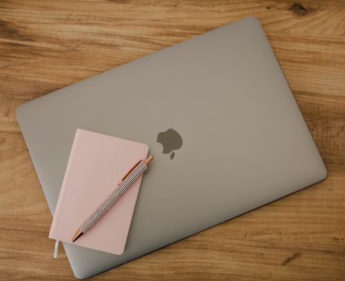 Free A sleek workspace featuring a laptop, pink notebook, and pen on a wooden desk. Stock Photo