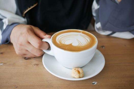 A hand holding a cappuccino with latte art and a cookie on a wooden table.
