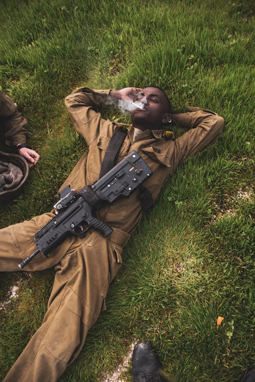 A Man in Brown Uniform Carrying Riffle Lying on a Grass Field while Smoking Cigarette
