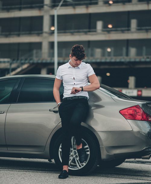 Free A man in casual attire leans on a car, checking his wristwatch in an urban setting. Stock Photo