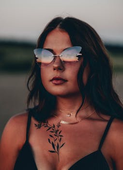 Woman with tattoos wearing butterfly sunglasses and tank top at sunset, showcasing bold style.