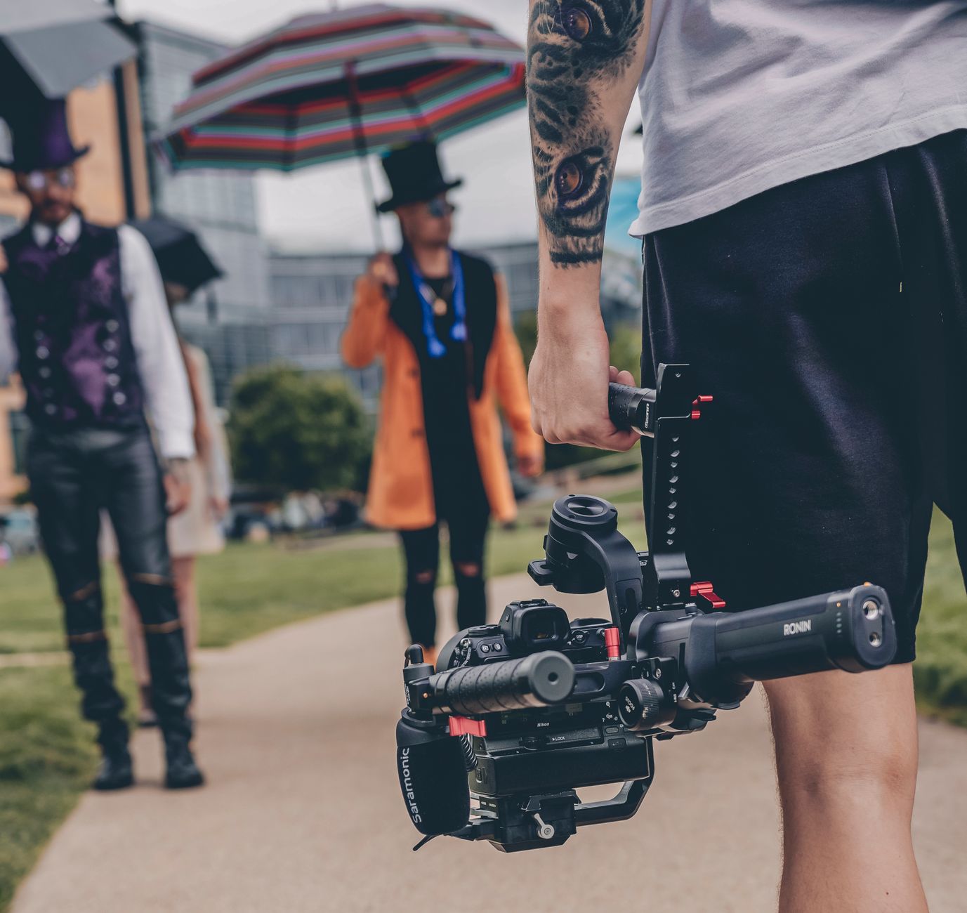 A tattooed videographer films people in cosplay costumes with a gimbal outdoors.