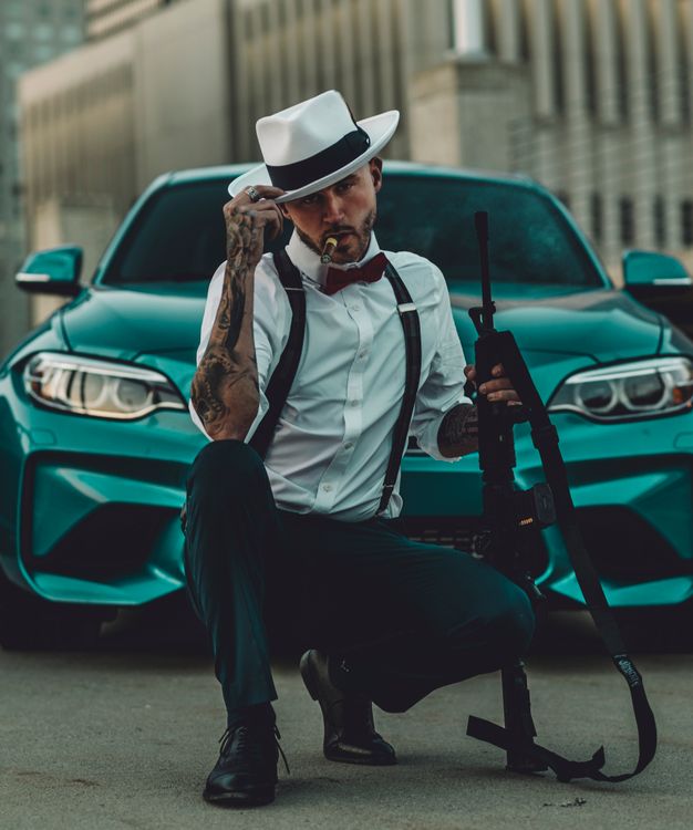 Free A man in 1920s gangster style poses with a rifle in front of a modern car. Stock Photo
