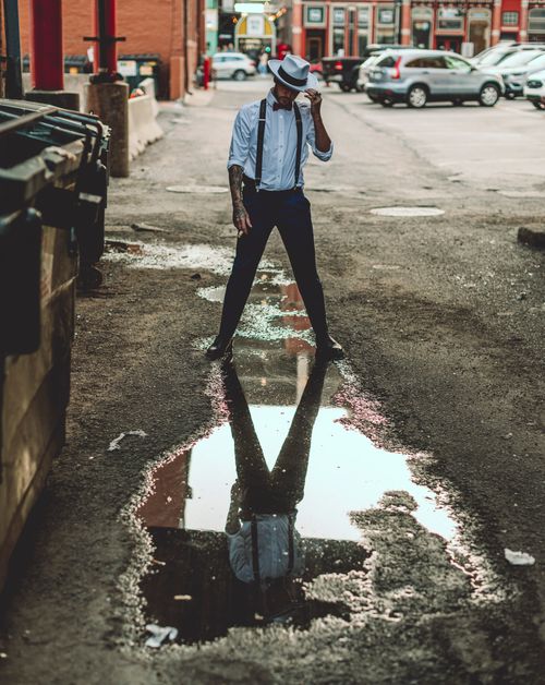 Free A fashionable man with a hat looks into a puddle in a gritty urban setting, creating a dynamic reflection. Stock Photo