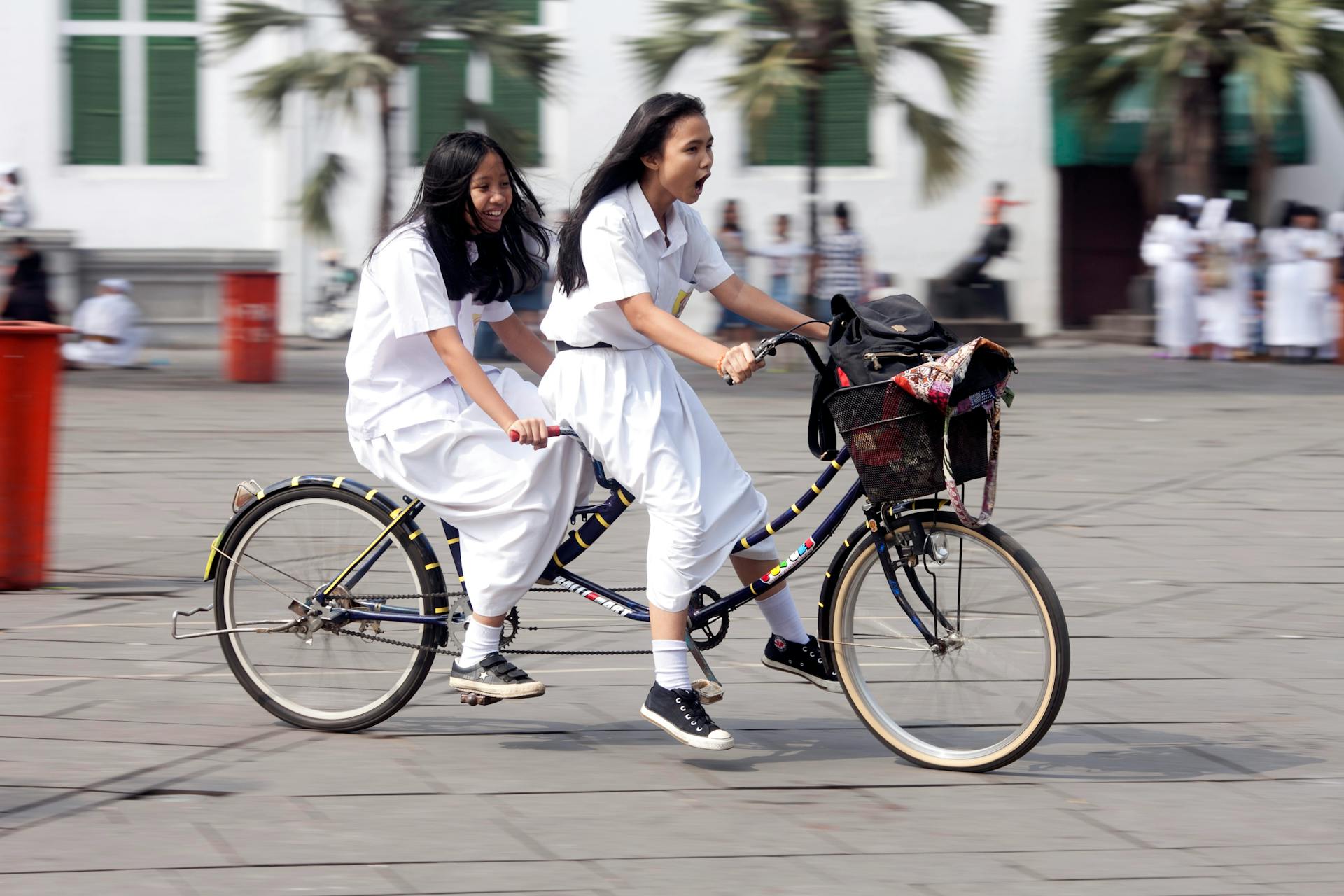 Two young girls in uniform enjoy a fun ride on a tandem bicycle in a lively outdoor setting.