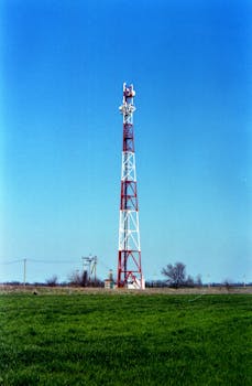 A red and white communication tower stands in a grassy field under a clear blue sky.