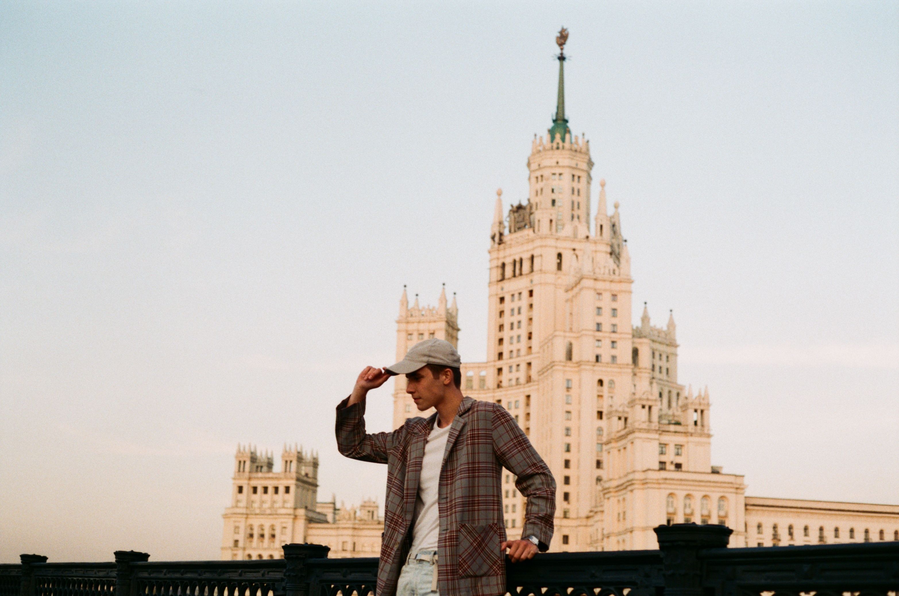 Young man in a plaid coat posing with a Moscow skyscraper in the background on a sunny day.