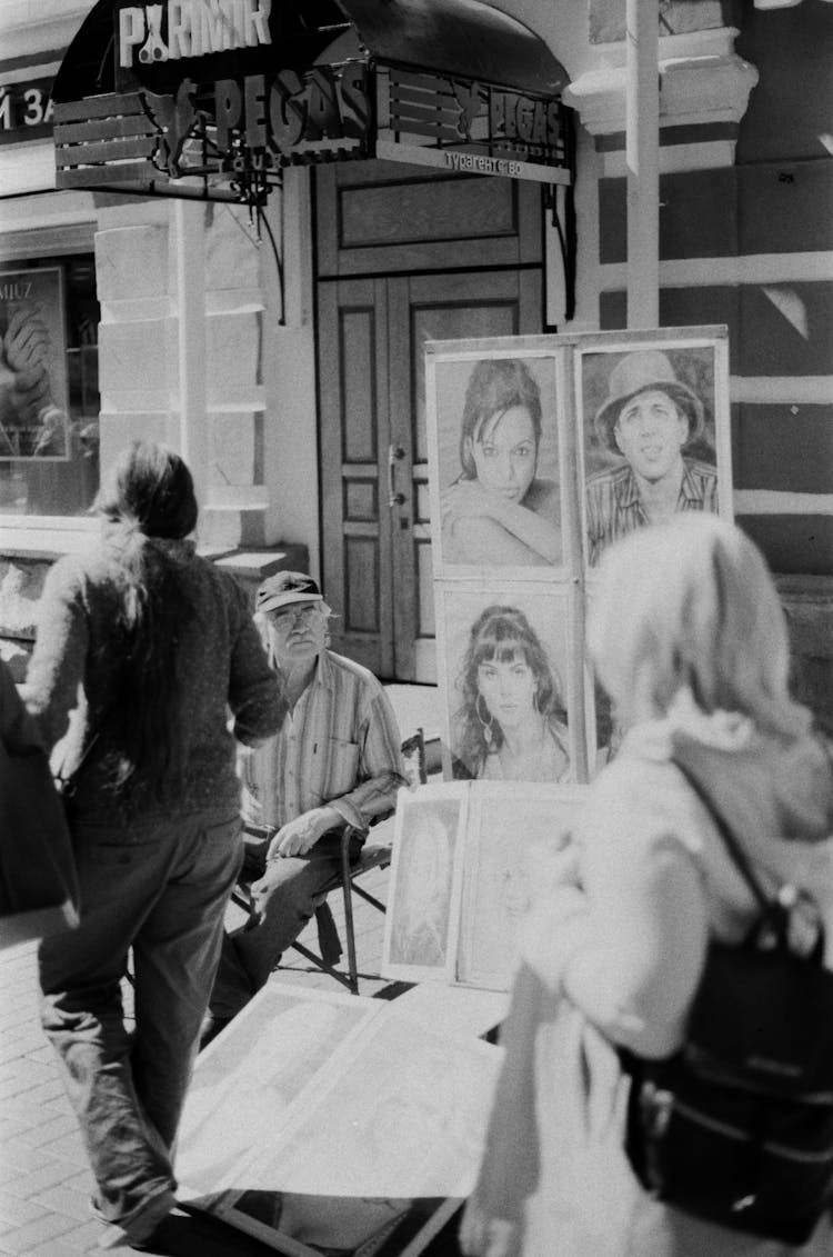 Grayscale Photography Of People Walking On The Street Near An Elderly Man Sitting Beside His Artwork