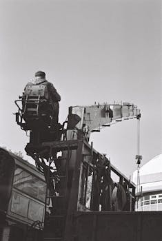 Black and white image of a crane operator working on site, showcasing industrial machinery.