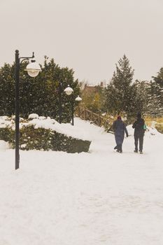 Two people walking through a snowy park in Istanbul, Türkiye, with street lamps and trees.