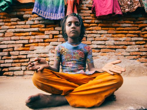Free Barefoot girl in colorful attire sits cross-legged against a rustic brick wall. Stock Photo