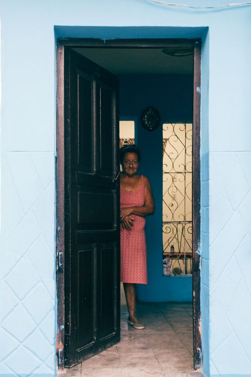 Free A woman in a pink dress stands at the open door of a blue house, smiling. Stock Photo