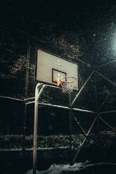A basketball hoop in winter as snowflakes softly fall, creating a serene night scene.
